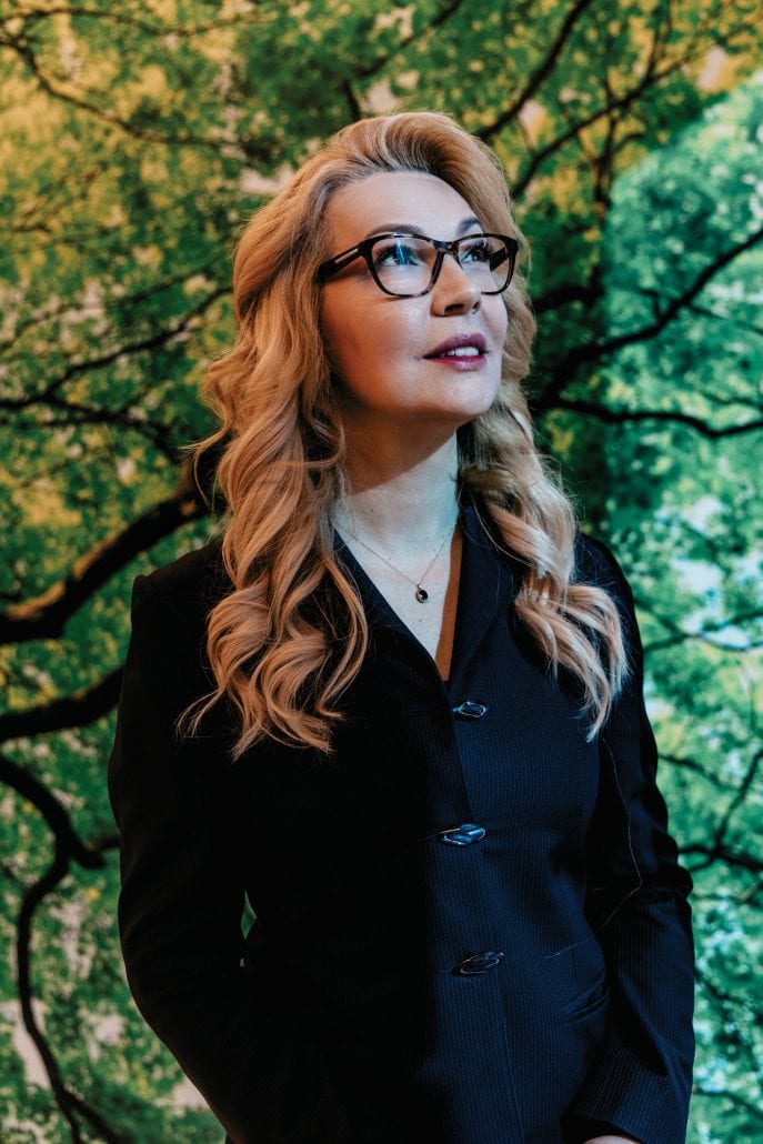 Woman in black stares up  with green trees behind her. 
