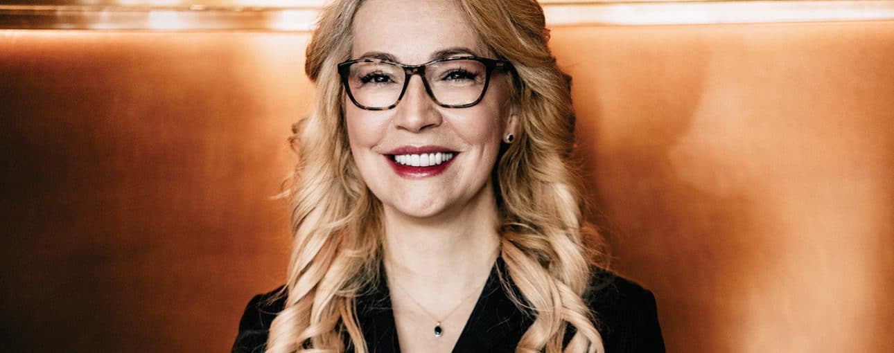 Woman with curled blond hair and glasses smiles at the camera.