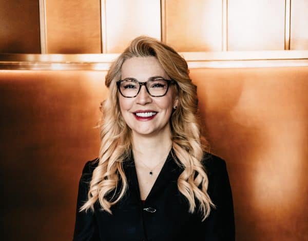 Woman with curled blond hair and glasses smiles at the camera.