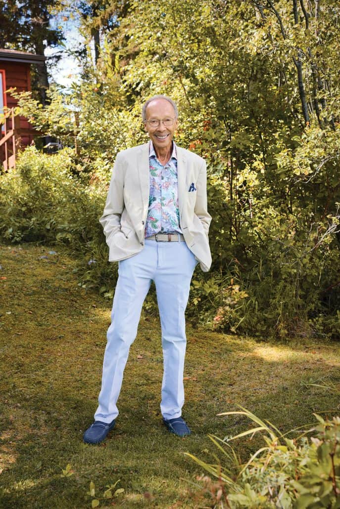 Man in a beige suit jacket standing with his hands in his pockets in front of bushes. 