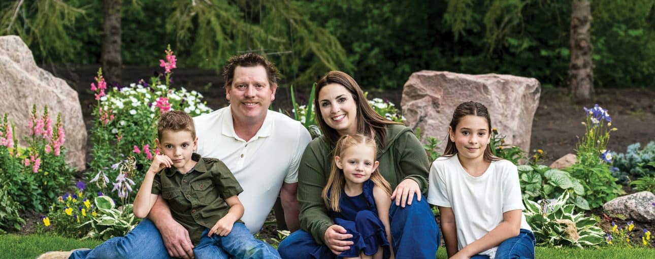 Family picture featuring mom, dad and three kids all sitting on the grass.