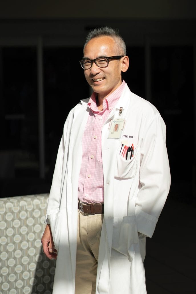 Man in a white lab coat and pink shirt looking at the camera while standing in sunlight.