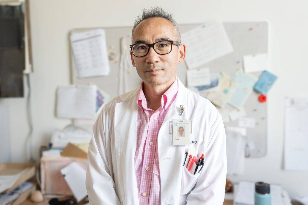 Man in a lab coat and pink shirt looking at the camera with papers on a bulletin board behind him.