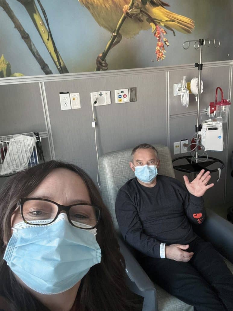 Women wearing a mask next to a man sitting and waving in a hospital chair. 