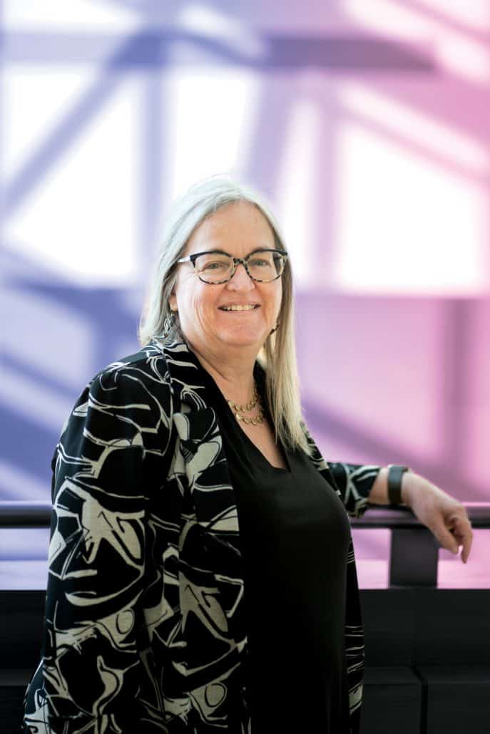 Woman smiles as she leans against a railing with a purple and pink background behind her. 