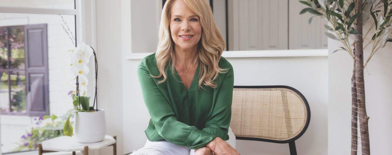 Woman in a green shirt and white pants sitting on a bench near the entryway of a home with an indoor plant next to her.