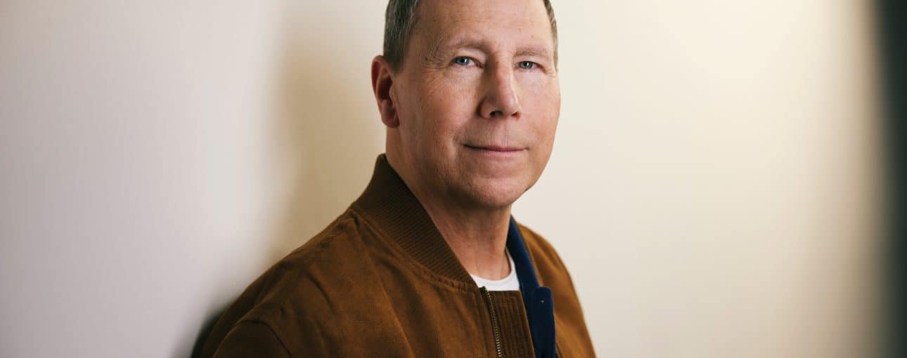 Man looks into the camera with beige wall behind him.