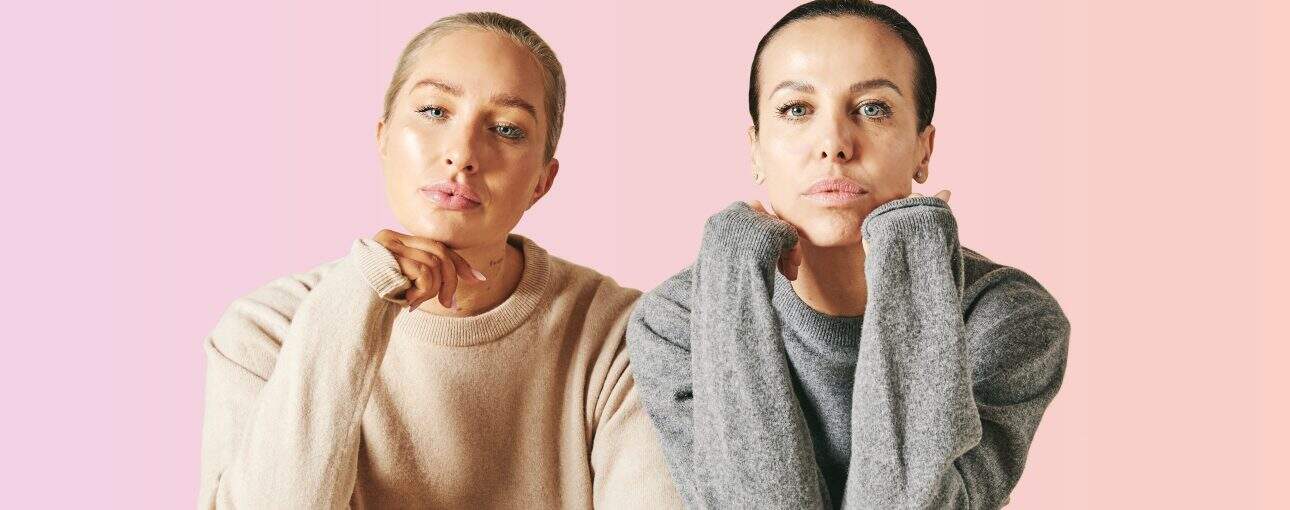 Two women pose looking into the camera with a pink background.