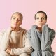 Two women pose looking into the camera with a pink background.