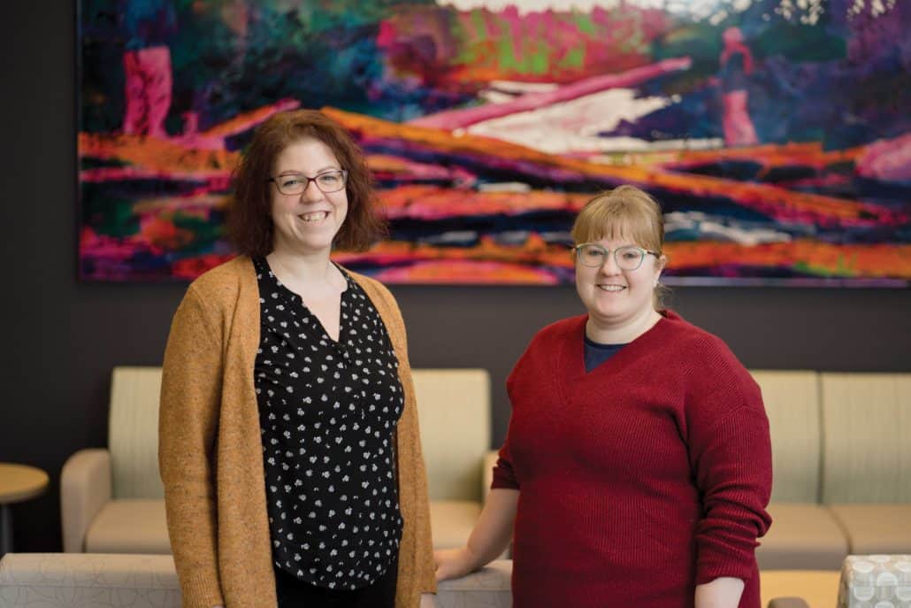 Two women smiling at the camera with a colourful painting in the background.