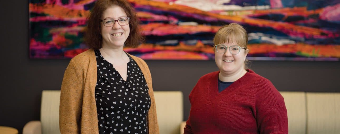 Two women smiling at the camera with a colourful painting in the background.