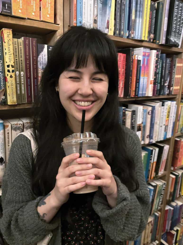 Woman smiling with a drink in her hand in front book shelves.