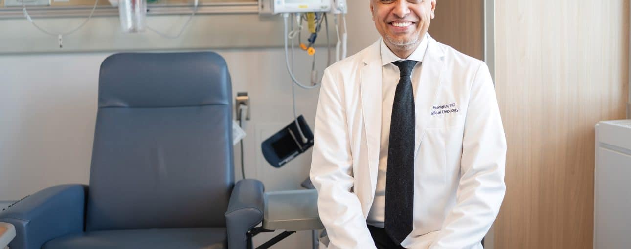 Doctor in a white coat smiles at the camera as he sits next to a patient chair.