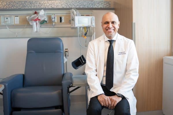 Doctor in a white coat smiles at the camera as he sits next to a patient chair.