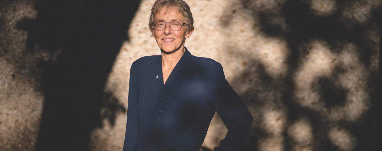 Woman standing in the sun with shadows from surrounding trees on the wall behind her.